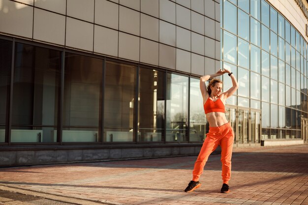 La donna atletica in abiti sportivi arancioni sta ballando, si sta scaldando all'aperto davanti all'edificio di vetro, copia spazio.