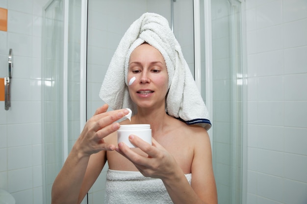 La donna attraente caucasica con un asciugamano bianco sulla testa applica una crema bianca sul viso dopo aver fatto la doccia o il bagno Trattamenti termali a casa