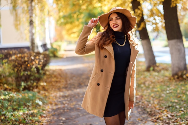 La donna attraente che porta il cappello a tesa larga e il cappotto beige sta trascorrendo il tempo all'aperto in autunno. Ritratto di donna alla moda nella stagione autunnale.