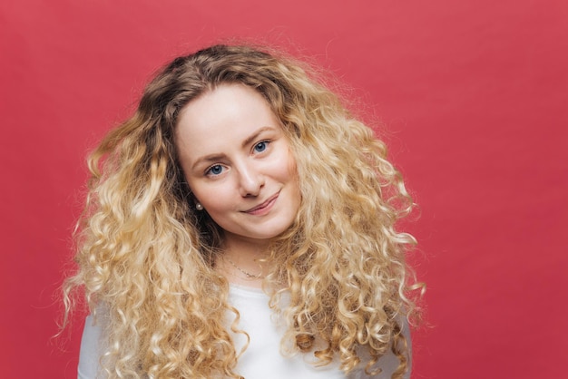 La donna attraente con i capelli ricci folti e gli occhi azzurri guarda positivamente la fotocamera si sente soddisfatta mentre incontra gli amici isolati su sfondo rosa brillante Concetto di bellezza ed emozioni della gente