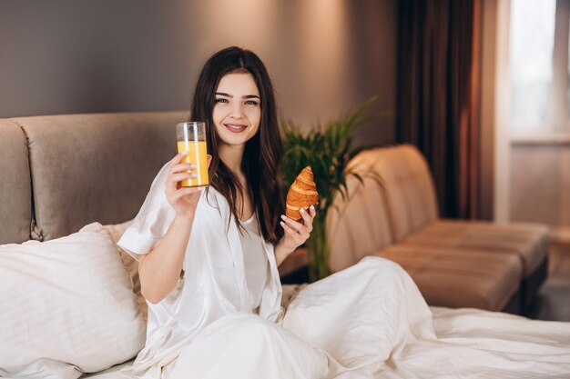 La donna beve il succo d'arancia a letto. colazione a letto. Una modella in pigiama bianco si siede sul letto e tiene in mano un bicchiere di succo d'arancia.