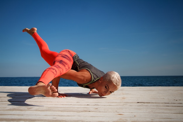 La donna bionda adulta con taglio di capelli corto pratica l'yoga sul pilastro contro lo sfondo del mare e del cielo blu