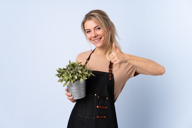 La donna bionda del giardiniere che tiene una pianta sopra la parete isolata che dà i pollici aumenta il gesto