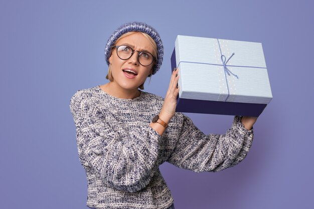 La donna bionda è in posa sorpresa mentre scuote un regalo su una parete blu dello studio con gli occhiali e il cappello