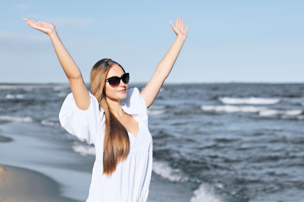 La donna bionda felice è sulla spiaggia dell'oceano in un abito bianco e occhiali da sole, alzando le mani.