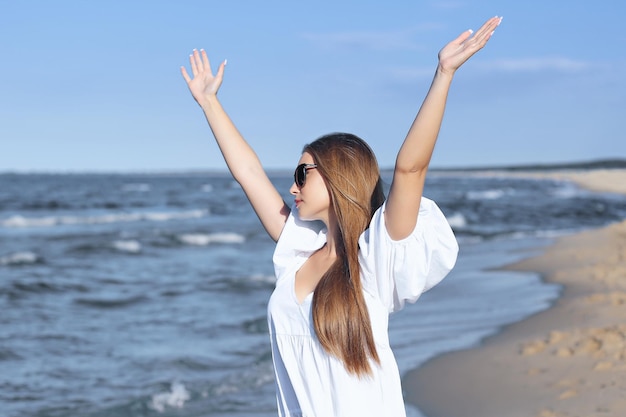 La donna bionda felice è sulla spiaggia dell'oceano in un abito bianco e occhiali da sole, alzando le mani