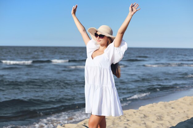 La donna bionda felice è sulla spiaggia dell'oceano in un abito bianco e occhiali da sole, alzando le mani.