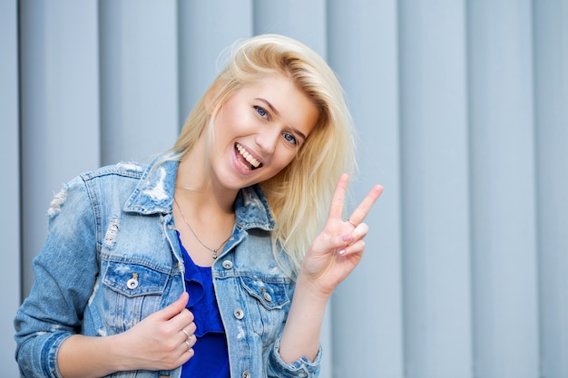 La donna bionda felice indossa una giacca di jeans che fa segno di pace con le dita. Spazio per il testo