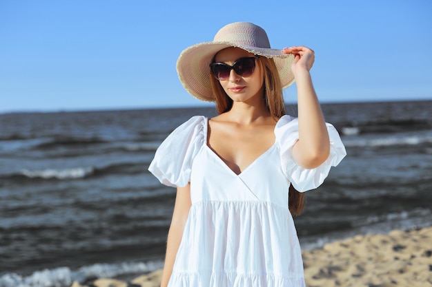 La donna bionda felice sta posando sulla spiaggia dell'oceano con gli occhiali da sole e un cappello. Sole della sera.