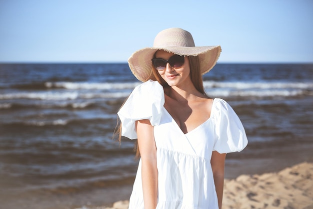 La donna bionda felice sta posando sulla spiaggia dell'oceano con occhiali da sole e un cappello. Sole serale