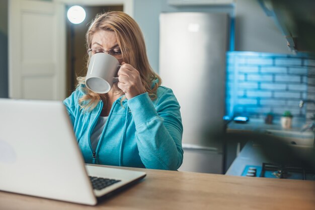 La donna bionda senior occupata sta bevendo una tazza di tè mentre lavora da casa al laptop in cucina