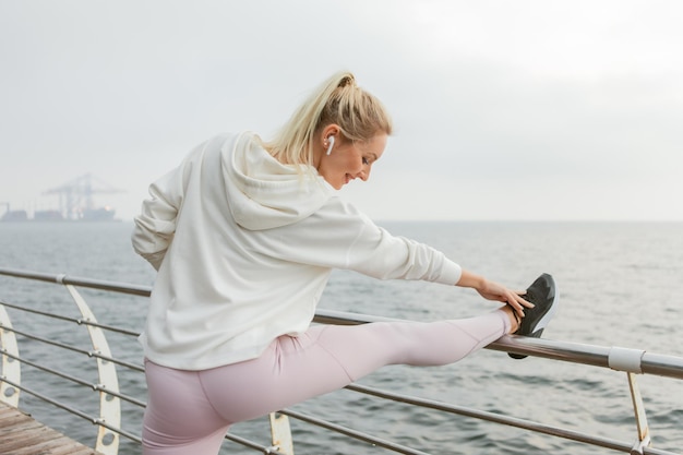 La donna bionda sexy slim fit sta praticando lo stretching delle gambe prima dell'allenamento all'aperto. Giovane ragazza vestita con felpa con cappuccio bianca si allena sulla spiaggia. Stile di vita sano, sport e fitness