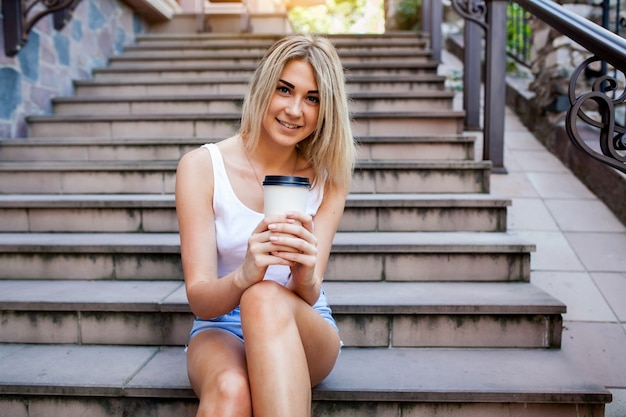 La donna bionda si siede sulle scale con un caffè