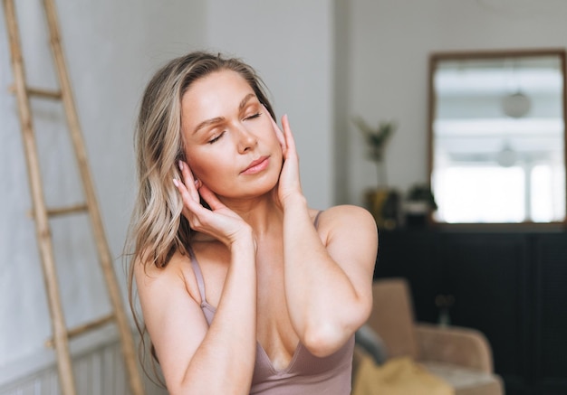 La donna bionda sorridente con i capelli lunghi fa il massaggio facciale in biancheria intima al bagno