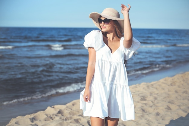 La donna bionda sorridente felice sta posando sulla spiaggia dell'oceano con occhiali da sole e un cappello