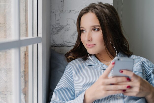 La donna bruna premurosa ascolta l'audiolibro tiene in mano lo smartphone moderno gode di un suono perfetto negli auricolari guarda pensieroso alla finestra vestita con abiti casual La donna ascolta la musica nella playlist