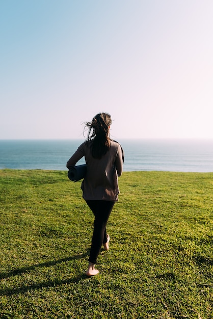 La donna cammina sulla schiena con una stuoia sull'erba per fare yoga di fronte al mare. Copia spazio
