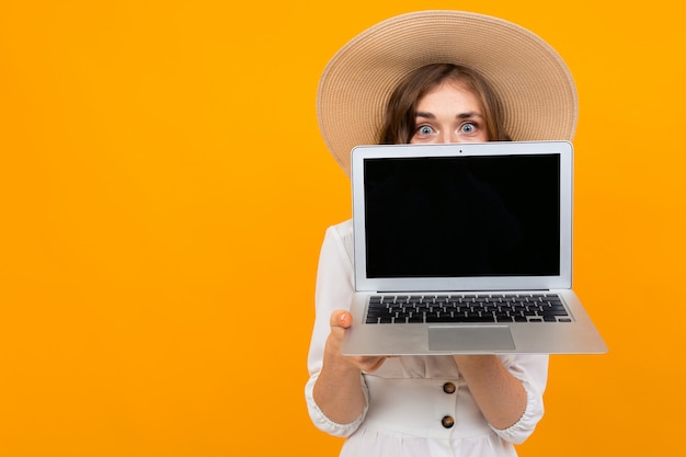 La donna castana allegra in un cappello tiene un computer portatile al livello del petto con lo schermo in avanti, ritratto su un'arancia