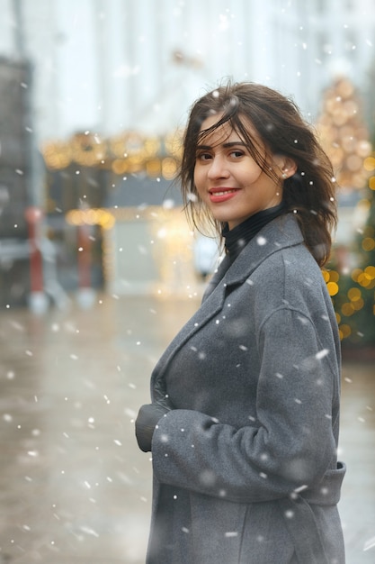 La donna castana allegra indossa un cappotto alla moda che cammina nella città decorata a Natale durante la nevicata