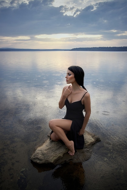 La donna castana di bellezza in un vestito nero posa in un lago del mare contro una priorità bassa del cielo blu. Donna sexy dai capelli lunghi e bellissimo trucco di bellezza sul viso