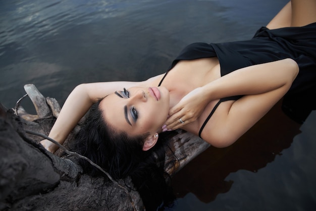 La donna castana di bellezza in un vestito nero posa in un lago del mare contro una priorità bassa del cielo blu. Donna sexy dai capelli lunghi e bellissimo trucco di bellezza sul viso