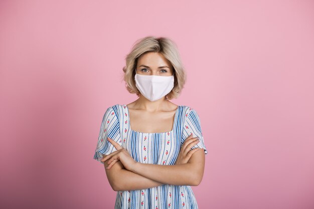 La donna caucasica bionda che indossa una maschera medica e un vestito sta esaminando la macchina fotografica su una parete dentellare dello studio