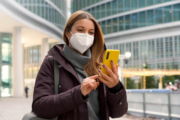 La donna caucasica con abiti invernali sta digitando con il dito chattando con qualcuno sullo smartphone mentre indossa una maschera protettiva all'esterno