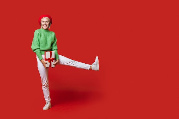 La donna caucasica con i capelli biondi che porta un cappello sta posando con un regalo su una parete rossa dello studio con qualcosa di pubblicità di spazio libero