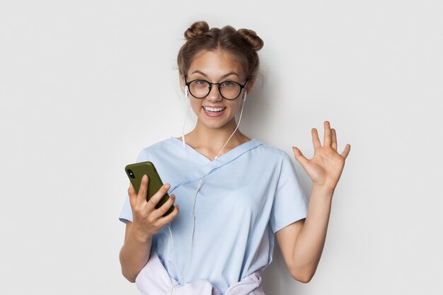 La donna caucasica con i capelli biondi sta sorridendo mentre ascolta la musica su un muro bianco usando gli auricolari e il telefono