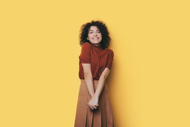 La donna caucasica con i capelli ricci sta sorridendo alla telecamera su un muro giallo che indossa un vestito alla moda