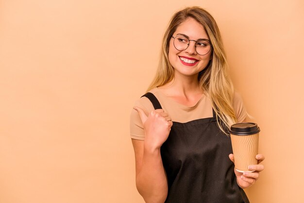 La donna caucasica del cameriere del ristorante che tiene un caffè da asporto isolato su fondo beige indica con il dito del pollice lontano ridendo e spensierato