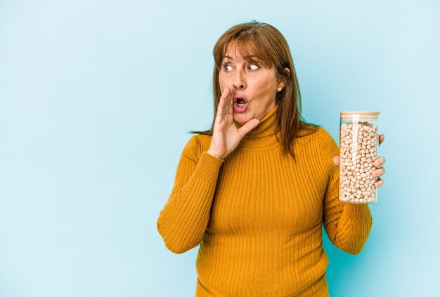 La donna caucasica di mezza età che tiene il barattolo di ceci isolato su sfondo blu sta dicendo una notizia segreta di frenata calda e sta guardando da parte