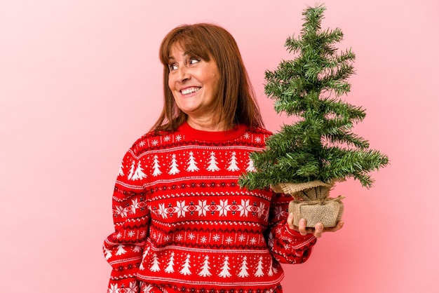 La donna caucasica di mezza età che tiene l'albero di Natale isolato su sfondo rosa guarda da parte sorridente, allegra e piacevole.