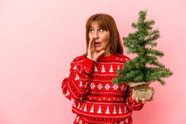 La donna caucasica di mezza età che tiene l'albero di Natale isolato su sfondo rosa sta dicendo una notizia segreta di frenata calda e sta guardando da parte