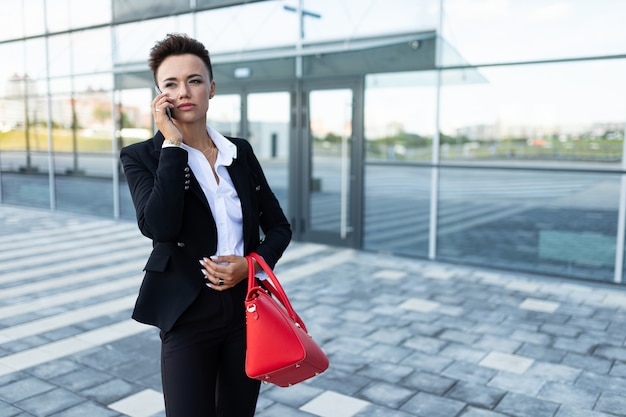 La donna caucasica in vestiti dell'ufficio e con la borsa rossa aspetta il collega vicino all'edificio per uffici
