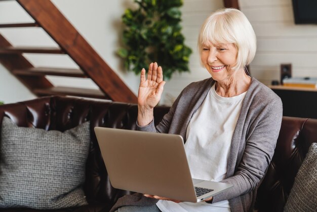 La donna caucasica senior sorridente che si siede sul colloquio dell'onda del divano durante la videochiamata sul computer portatile