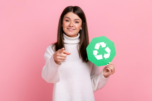 La donna che tiene il cartello di riciclaggio verde, indicando la fotocamera, invita a salvare il nostro pianeta dall'inquinamento, indossando un maglione bianco in stile casual. Studio indoor girato isolato su sfondo rosa.