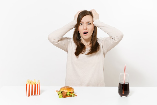 La donna colpita ha messo le mani sulla testa alla tavola con la cola delle patate fritte dell'hamburger in bottiglia di vetro isolata su fondo bianco. Una corretta alimentazione o un classico fast food americano. Area pubblicitaria con copia spazio.