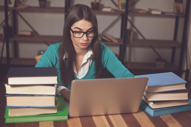 La donna con gli occhiali con un sacco di libri e laptop è seduta al tavolo di legno