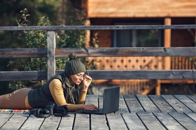 La donna con gli occhiali si trova sulla terrazza in legno e lavora sul computer portatile. concetto di calore autunnale, atmosfera e comfort, lavoro nella natura, freelance