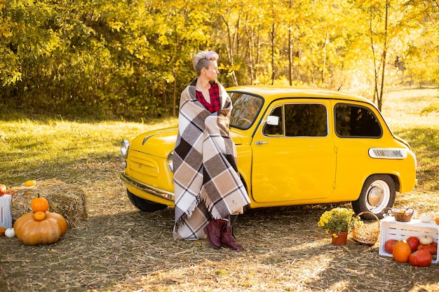 La donna con i capelli corti avvolta in una coperta vicino alla retro automobile gialla in autunno fiorisce lo sfondo con alberi dorati