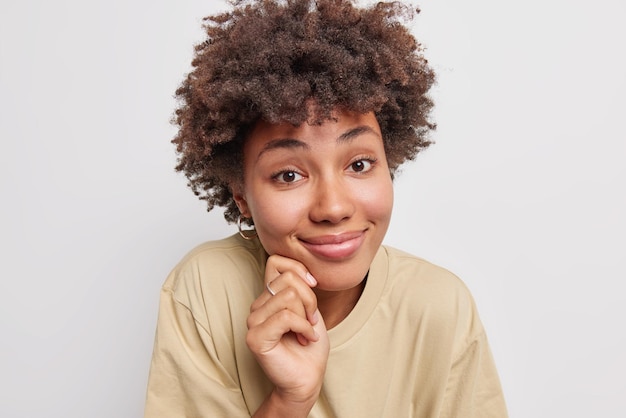 la donna con i capelli ricci tocca il viso e sorride piacevolmente sembra ha un'espressione soddisfatta indossa una maglietta casual isolata su bianco ascolta qualcosa di buono