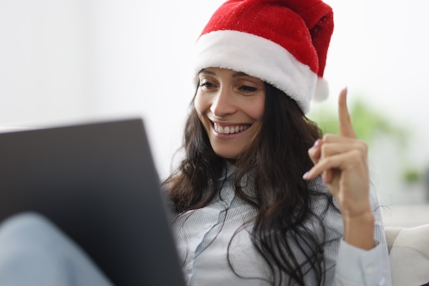 La donna con i sorrisi del cappello di Babbo Natale tiene i pollici in su e guarda nel computer portatile.