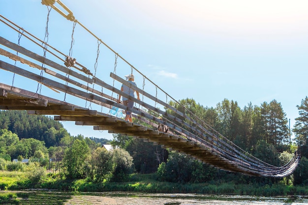 La donna con il cane cammina lungo il vecchio ponte sospeso in legno sul fiume in una giornata di sole estivo. Paesaggio naturale. Cane sul ponte.