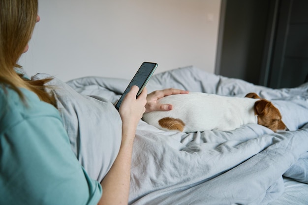 La donna con il cane che si rilassa sul letto usa lo smartphone