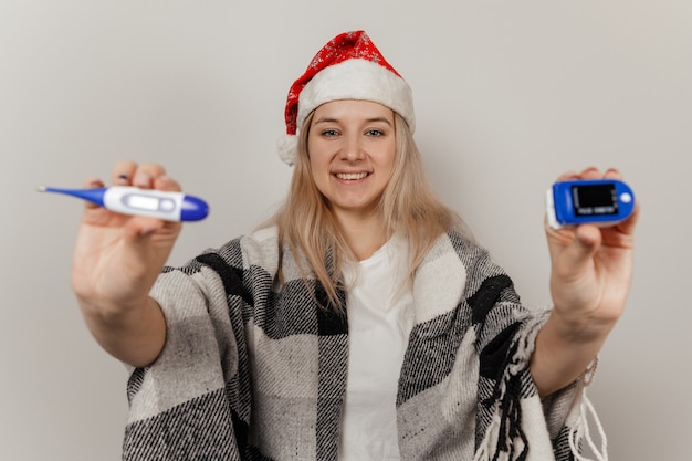 La donna con il cappello da Babbo Natale e l'involucro tiene il termometro e il pulsossimetro su sfondo grigio isolato