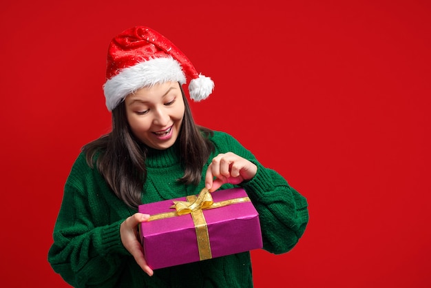 La donna con un cappello di Natale scompatta un regalo. Su uno sfondo rosso.