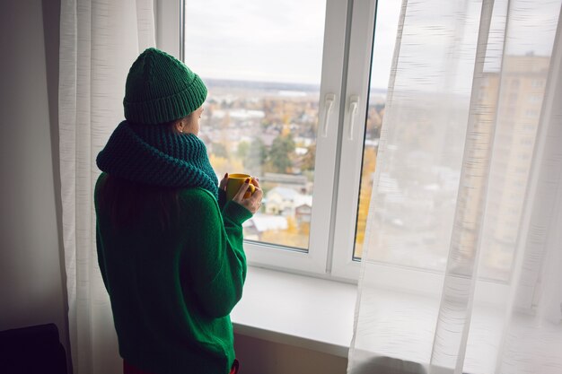 La donna con un maglione lavorato a maglia verde e un cappello si trova alla finestra della casa con una tazza di caffè in autunno e guarda in lontananza la foresta