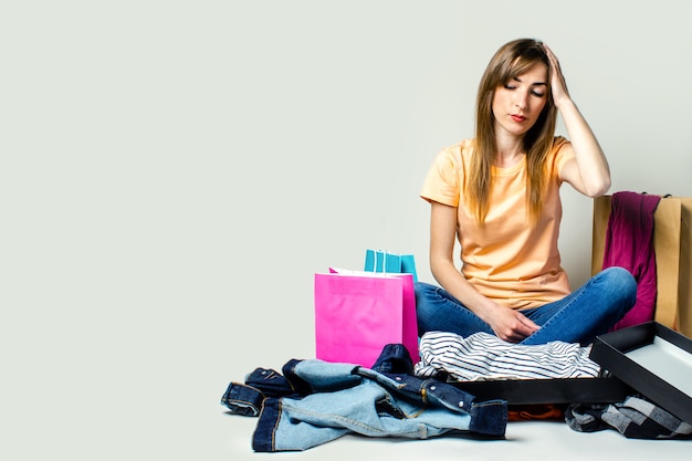 La donna con una faccia triste si siede sul pavimento circondato da diversi abiti e borse della spesa