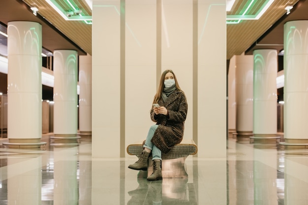 La donna con una maschera medica per evitare la diffusione di COVID è seduta con uno smartphone e aspetta un treno della metropolitana. Una ragazza con i capelli lunghi e una mascherina chirurgica sta mantenendo le distanze sociali in metropolitana.